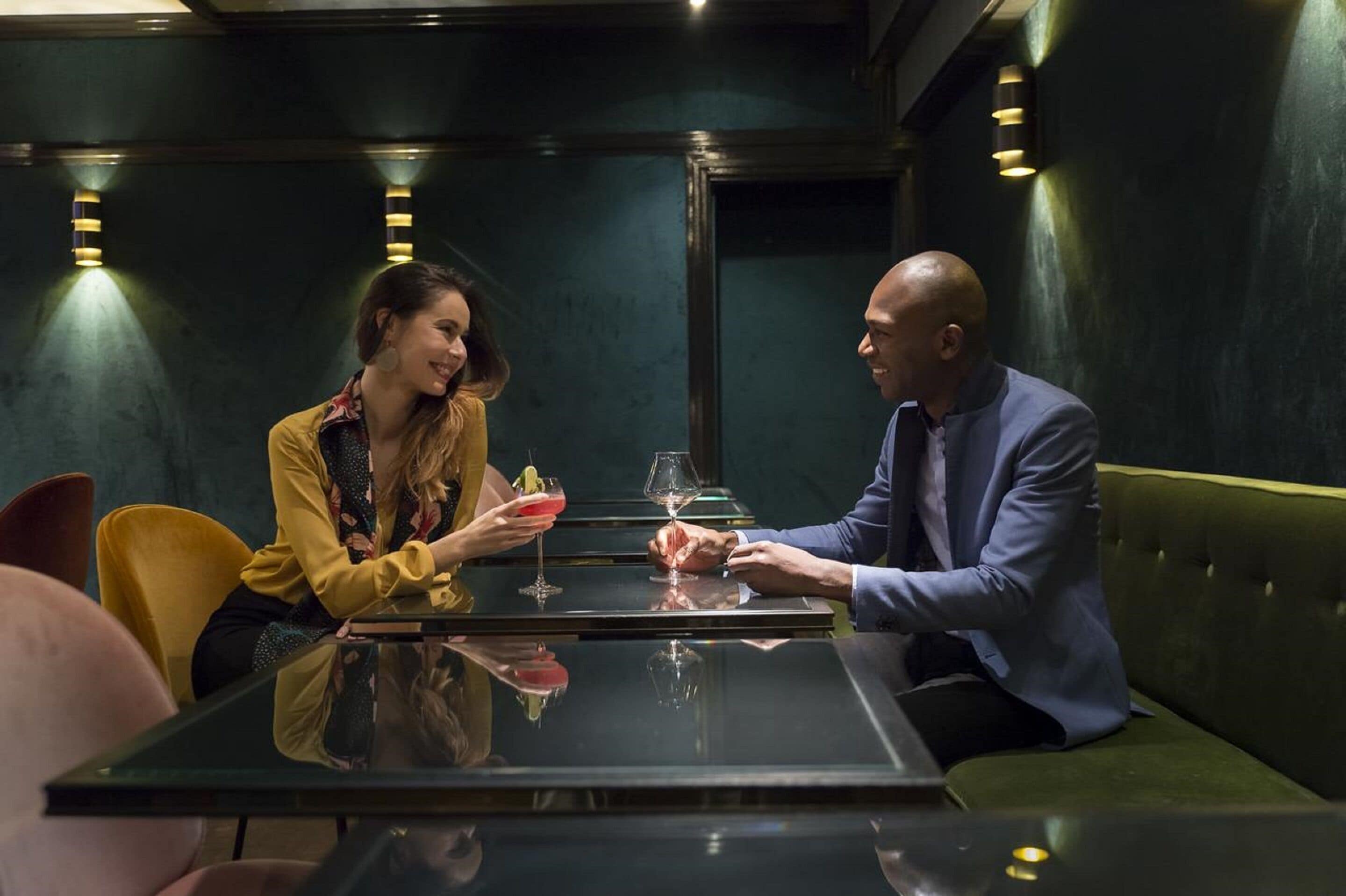 A couple seated at a table with drinks, in a room with dark green walls and modern lighting.