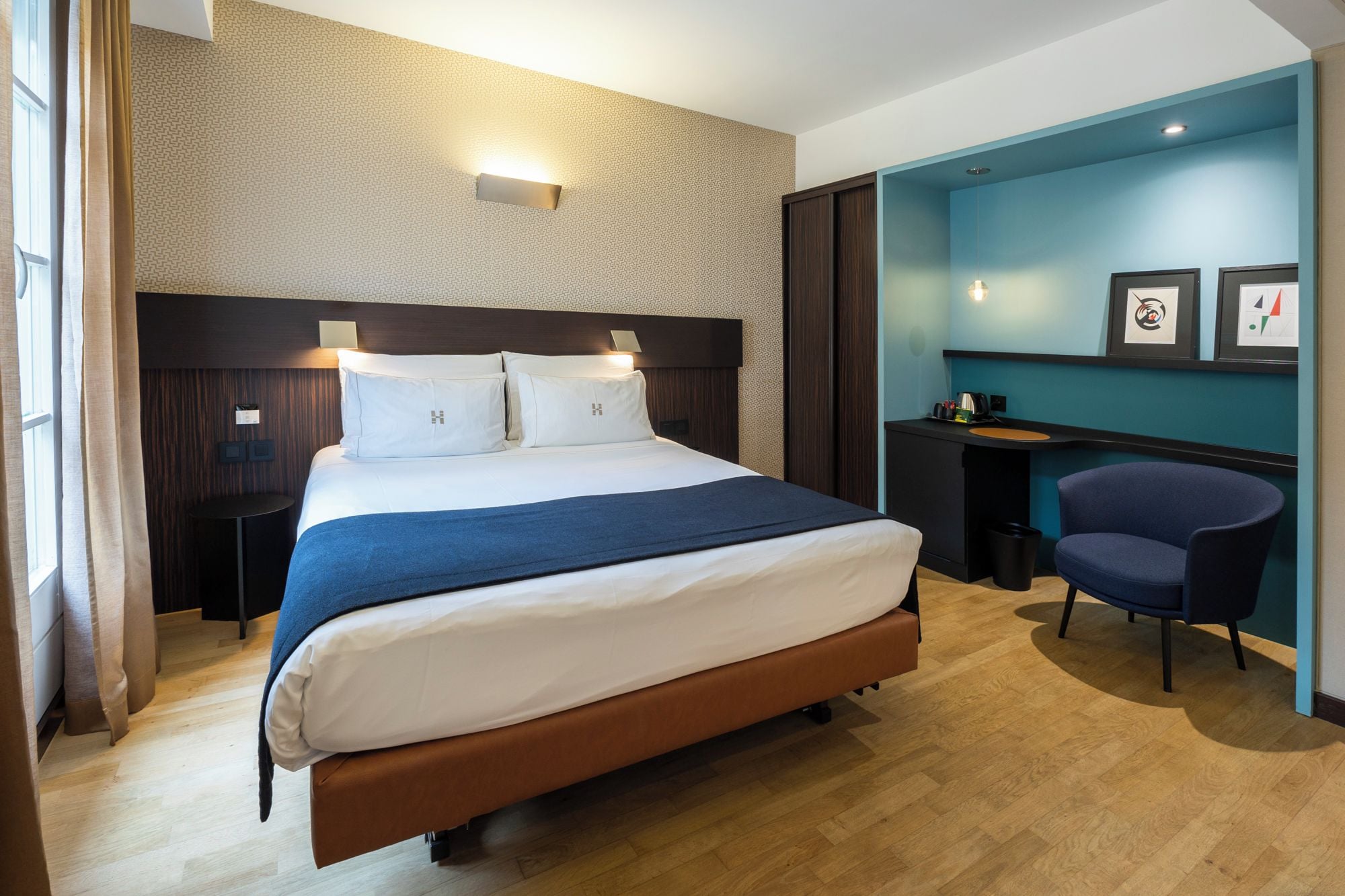 A hotel room with a large bed, a desk, a chair, and a wall with framed pictures.