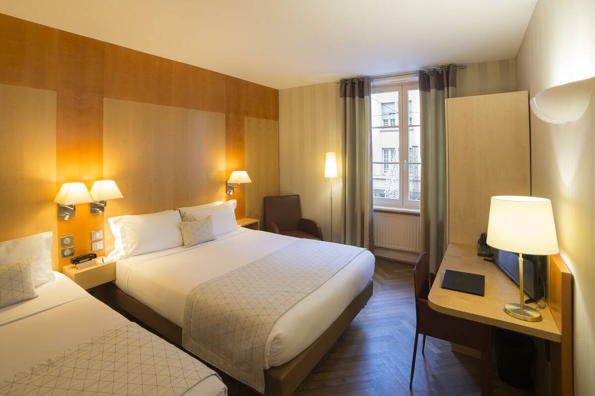 A hotel room with two beds, a desk with a chair, and a large window with curtains.