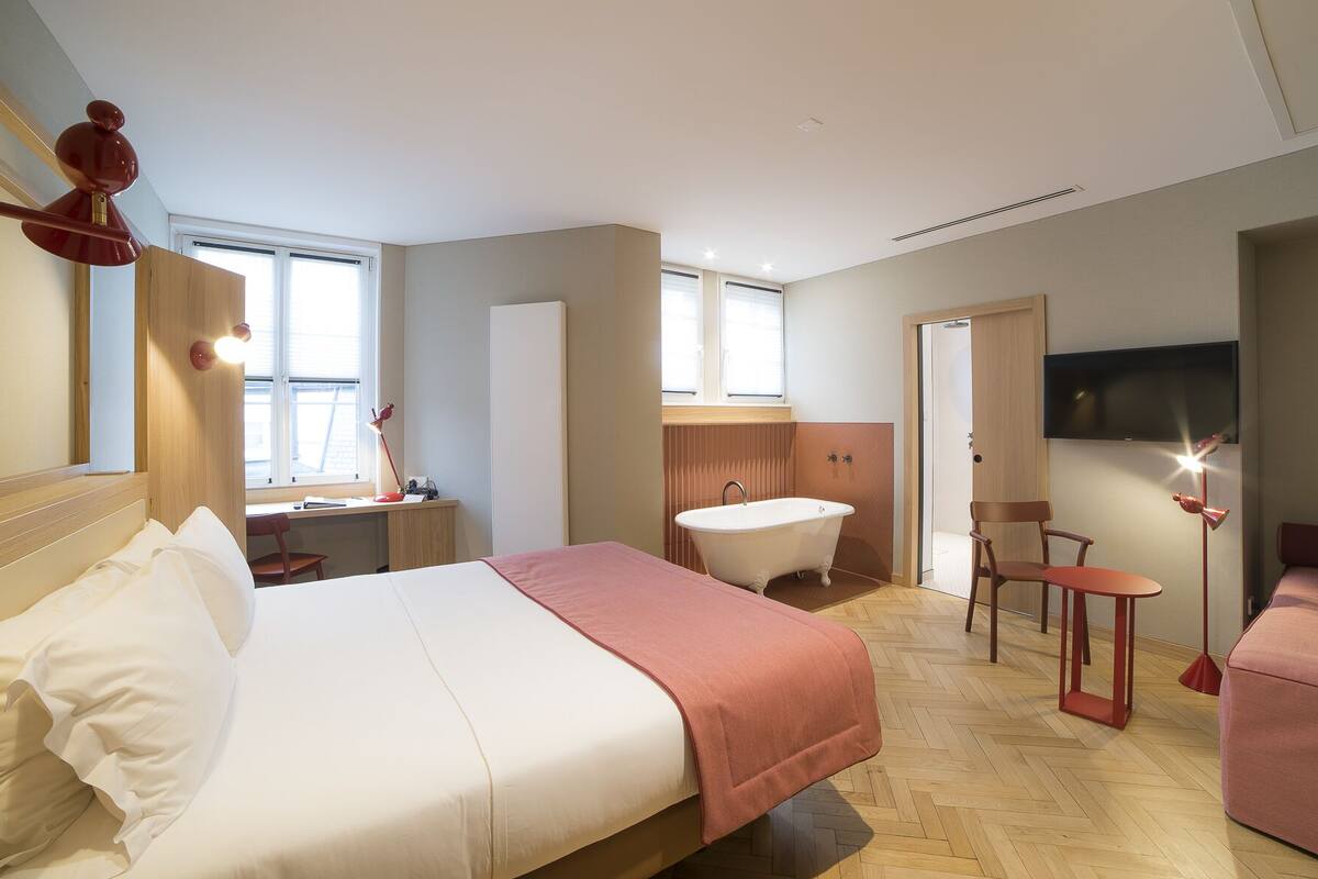A modern hotel room with a large bed, a sitting area with a chair and a small table, a bathtub, and a television mounted on the wall.