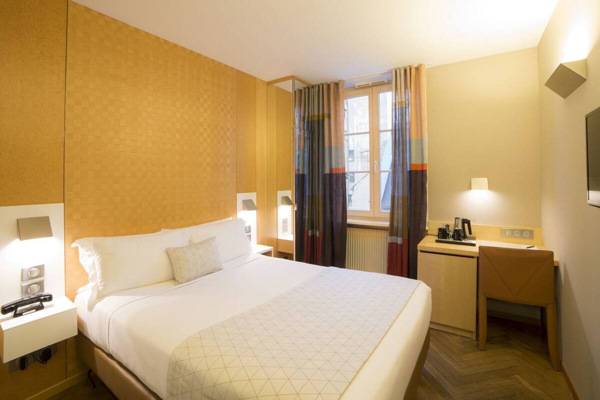 A hotel room with a bed, bedside tables, a desk, a chair, a television, and a window with curtains.