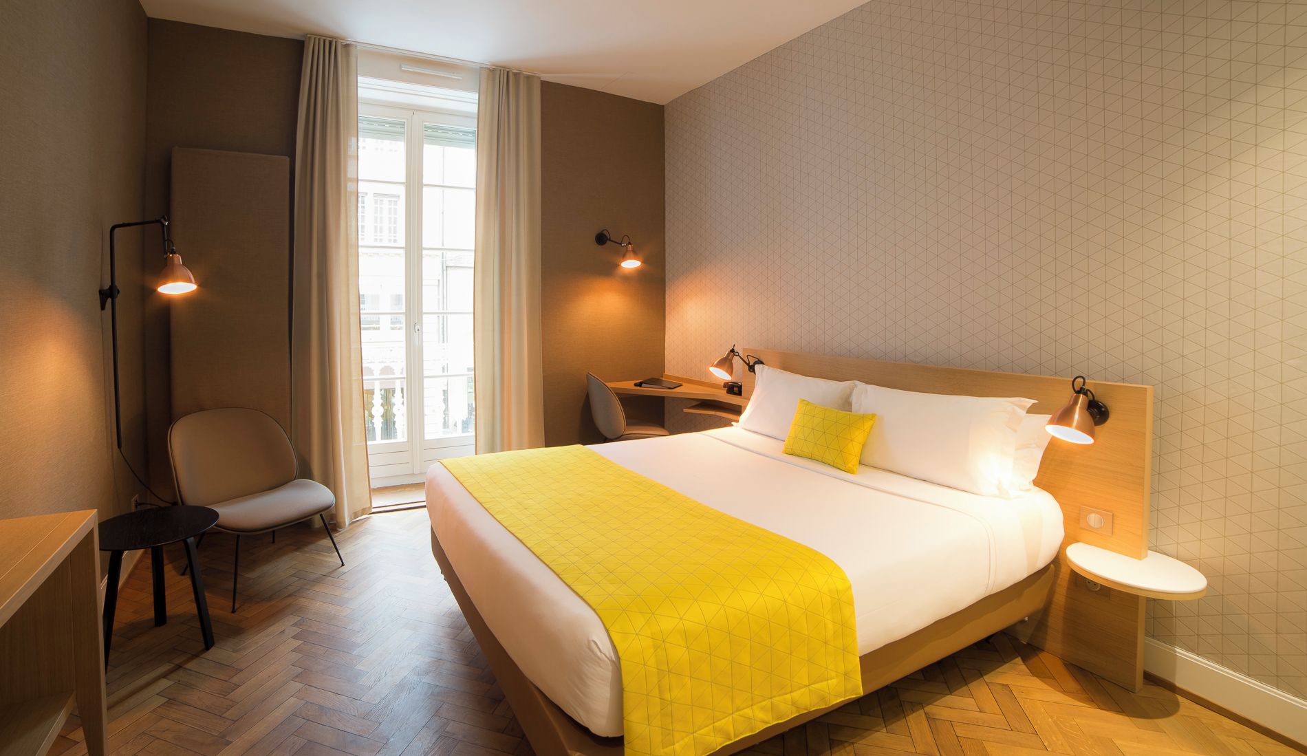 Quarto de hotel com cama, escrivaninha, cadeira, mesa de cabeceira e abajur.
