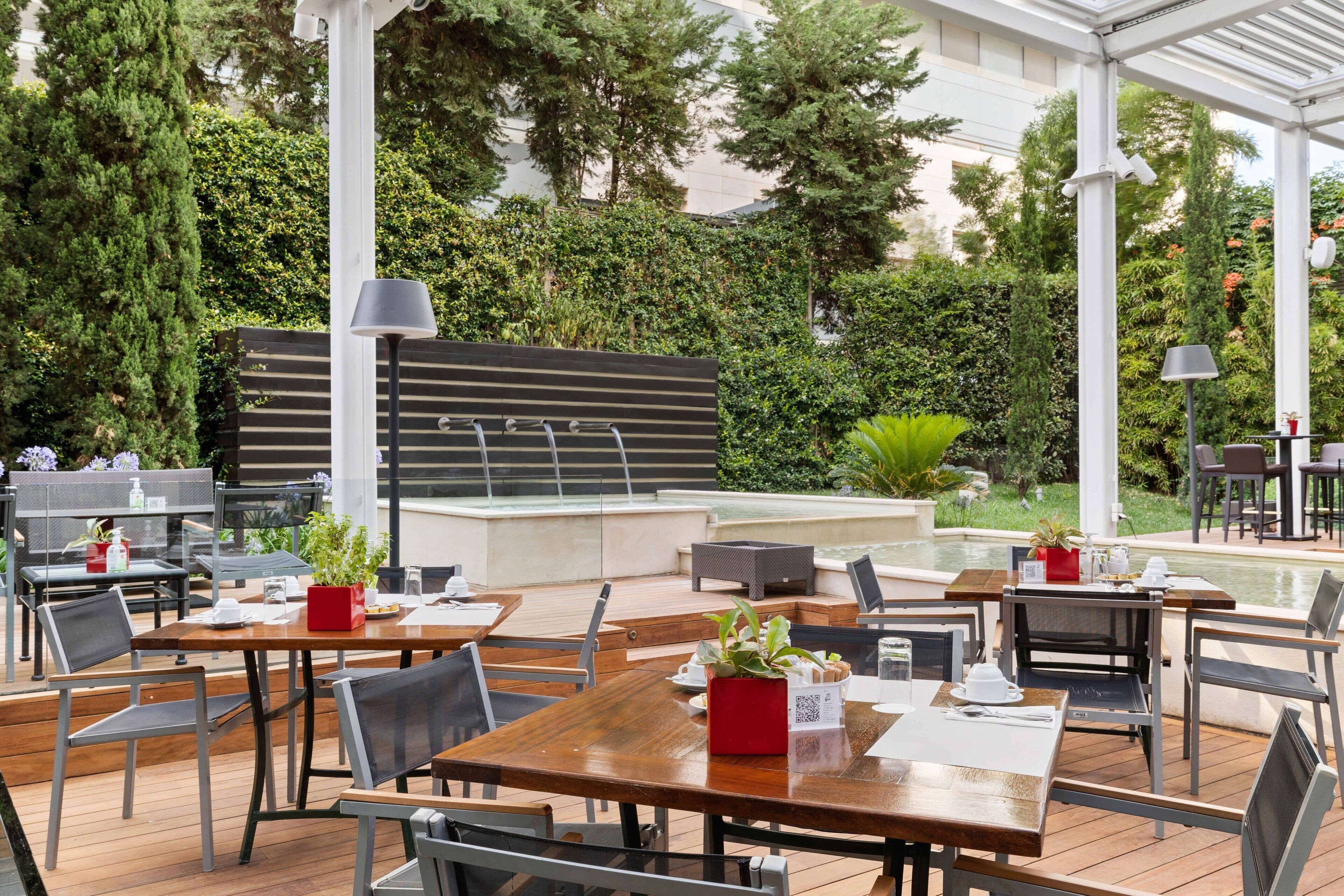 Un área de comedor al aire libre con mesas y sillas, una fuente de agua y abundante vegetación.