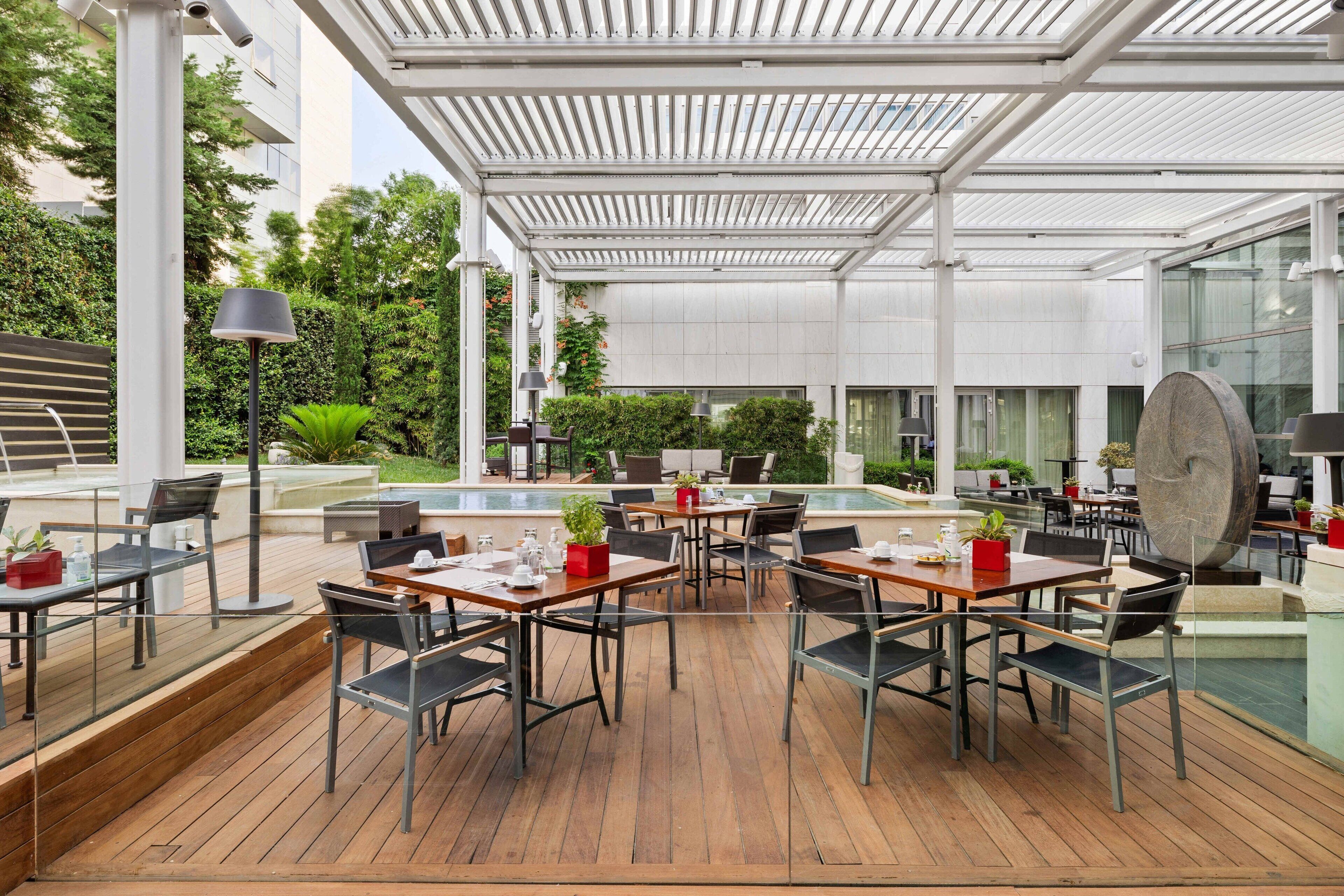 An outdoor dining area with wooden tables and chairs, surrounded by a glass enclosure, and a pool in the background.