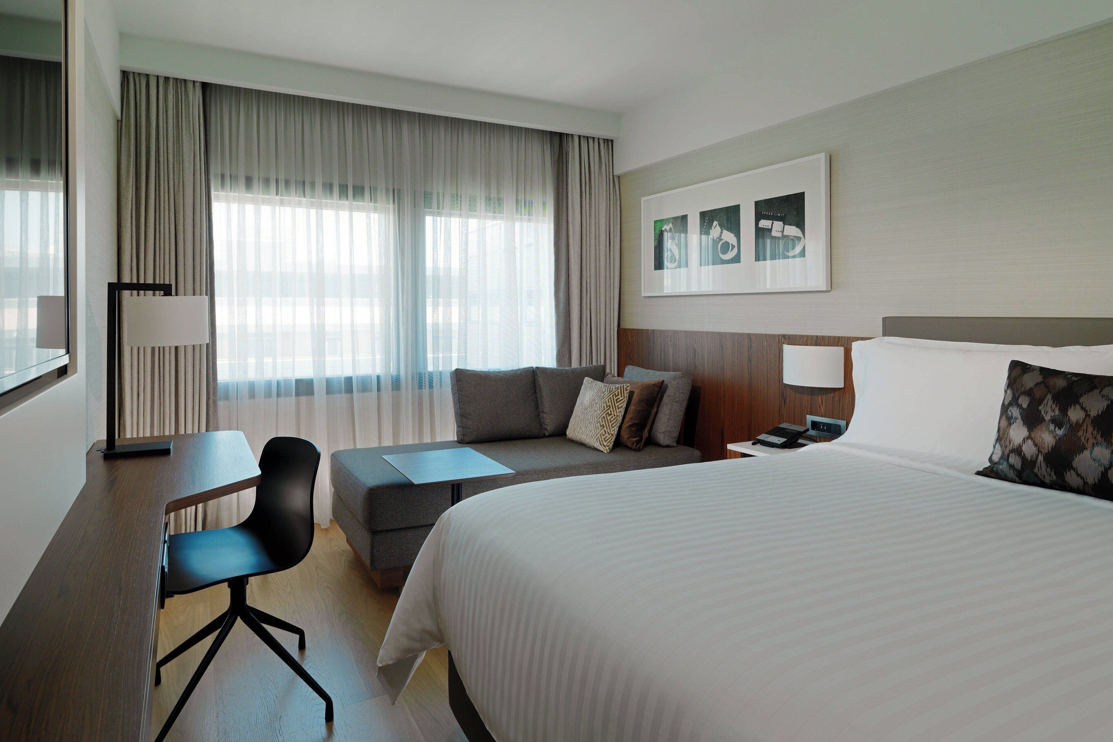 A hotel room with a large bed, a sofa, a desk, and a chair. There is a window with curtains, a wall-mounted lamp, and a framed picture on the wall.