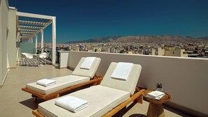 Terraza en la azotea con dos tumbonas, una mesita con un libro y vistas a un paisaje urbano y montañas al fondo.