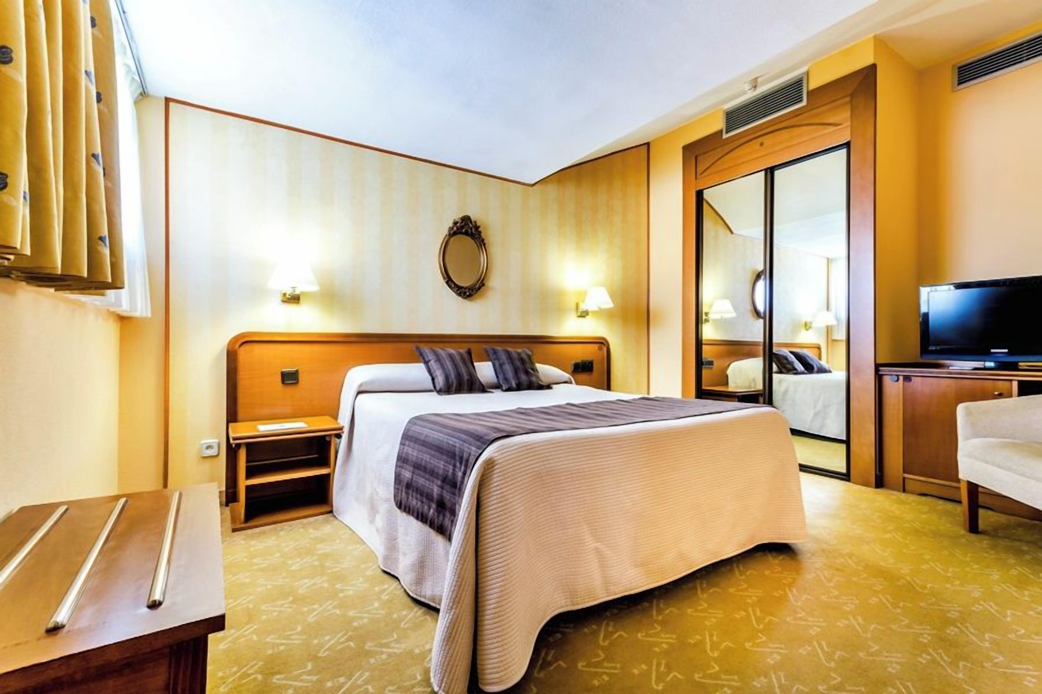 Una habitación de hotel con cama, mesitas de noche, televisión y espejo.