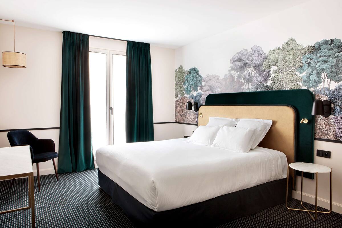 A hotel room with a large bed, a desk, a chair, and a view of trees through a window.