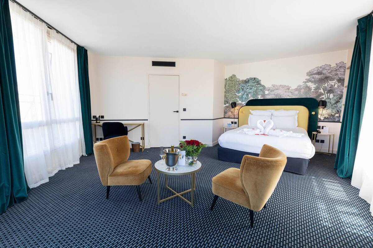 A hotel room with a bed, two armchairs, a small round table with a vase of flowers, and a desk.