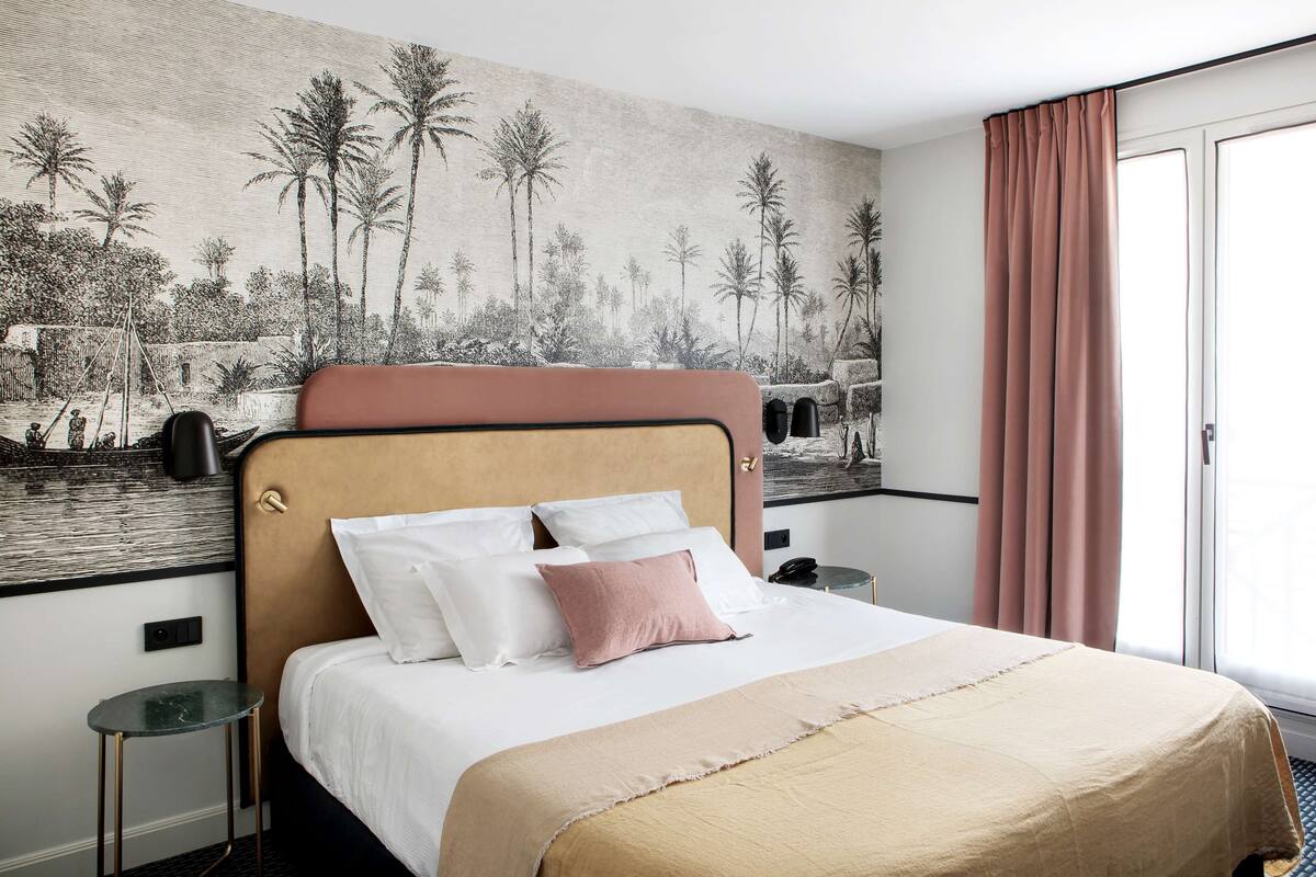 A hotel room with a bed, a nightstand, a wall mural, and a window with curtains.