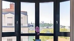 Quarto com uma janela grande, duas cadeiras, uma mesa pequena com um vaso de flores e vista para edifícios.