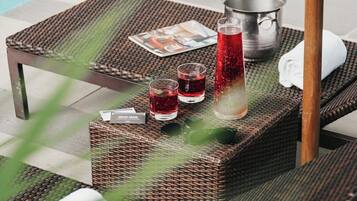 Un área de descanso junto a la piscina con mobiliario de mimbre, un cubo de metal y dos vasos con bebida roja.
