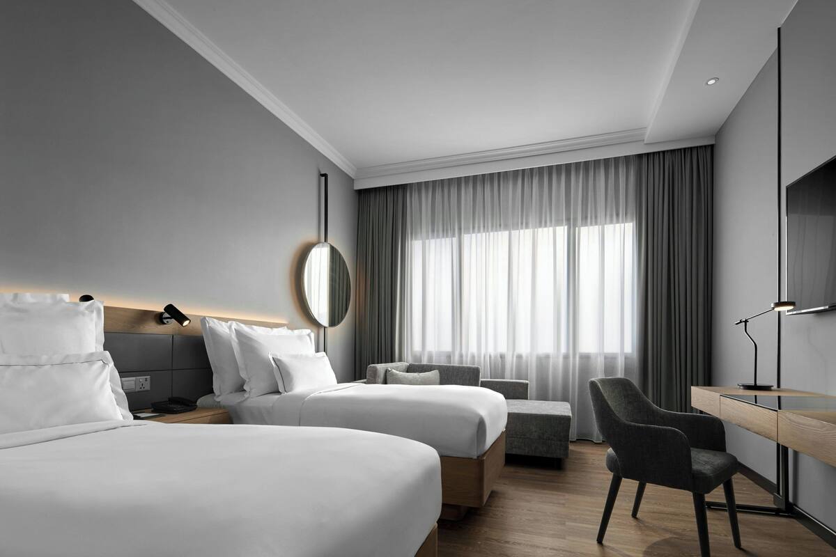 Een moderne hotelkamer met twee bedden, een bureau, een stoel en een groot raam met gordijnen.