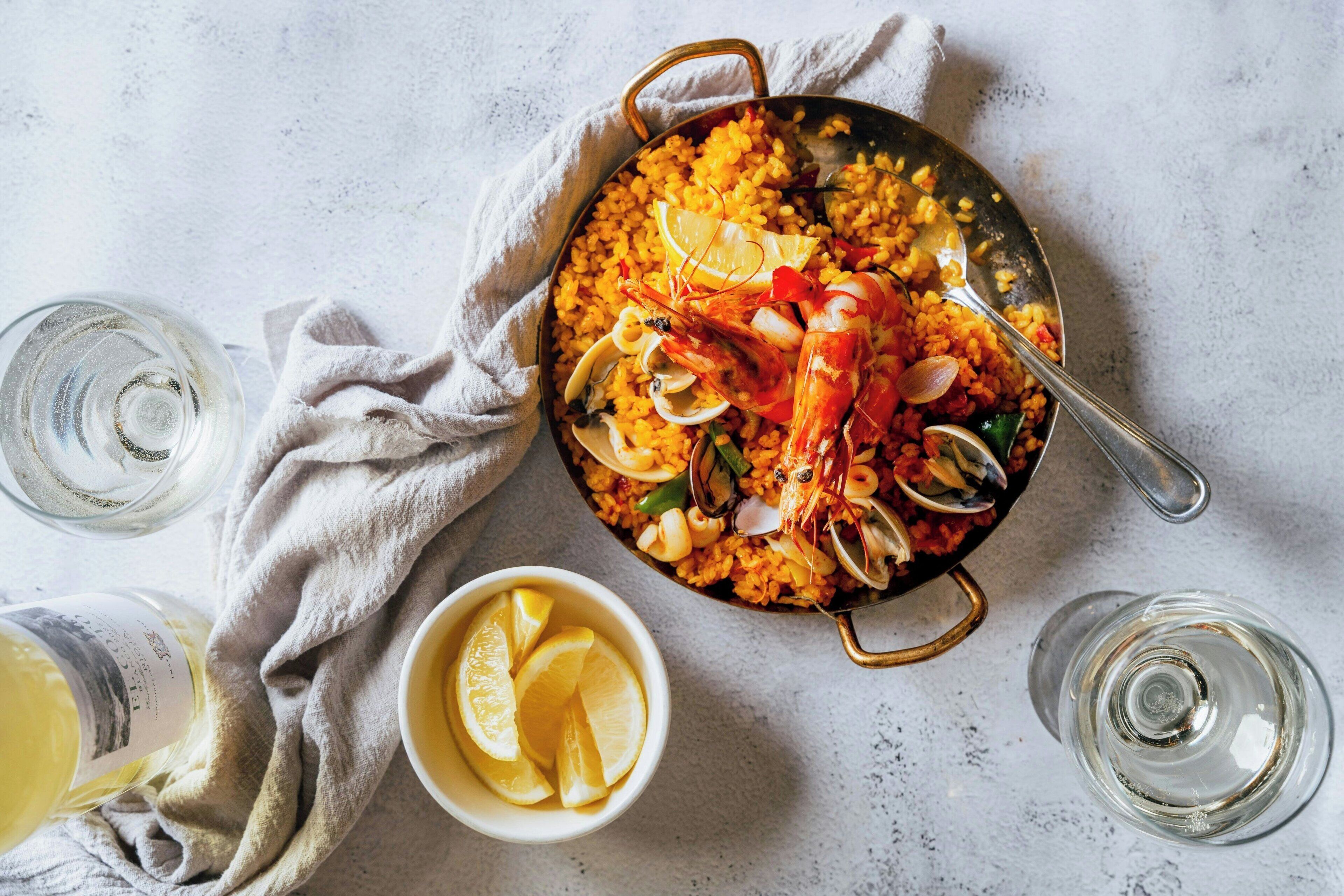 Eine Pfanne mit Meeresfrüchte-Paella, mit Zitronenspalten garniert, begleitet von Gläsern Weißwein und einer Schale Zitronenscheiben.