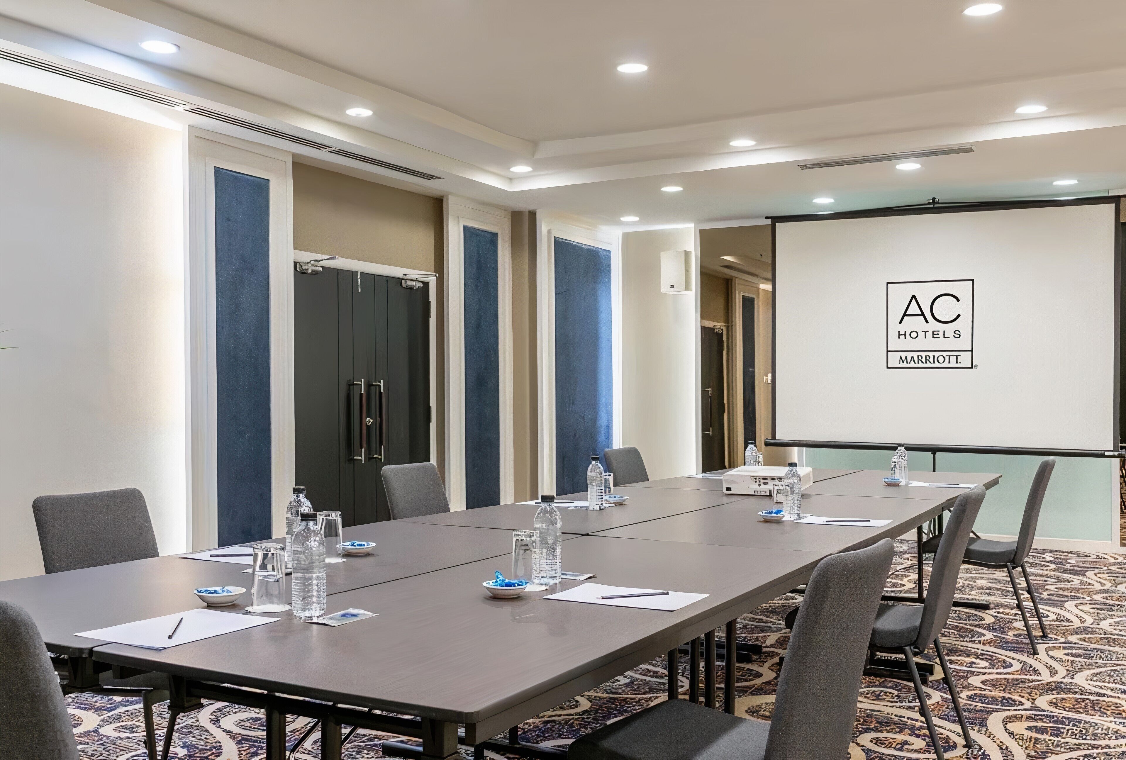 Ein Konferenzraum mit langem Tisch, Stühlen, Wasserflaschen und einer Projektionsfläche, auf der das Logo von AC Hotels Marriott zu sehen ist.