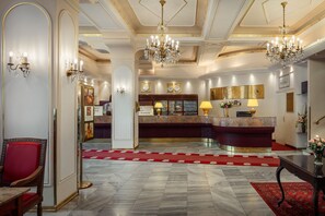 Un hall d’entrée luxueux avec un sol en marbre, un comptoir d’accueil, un tapis rouge et un lustre.