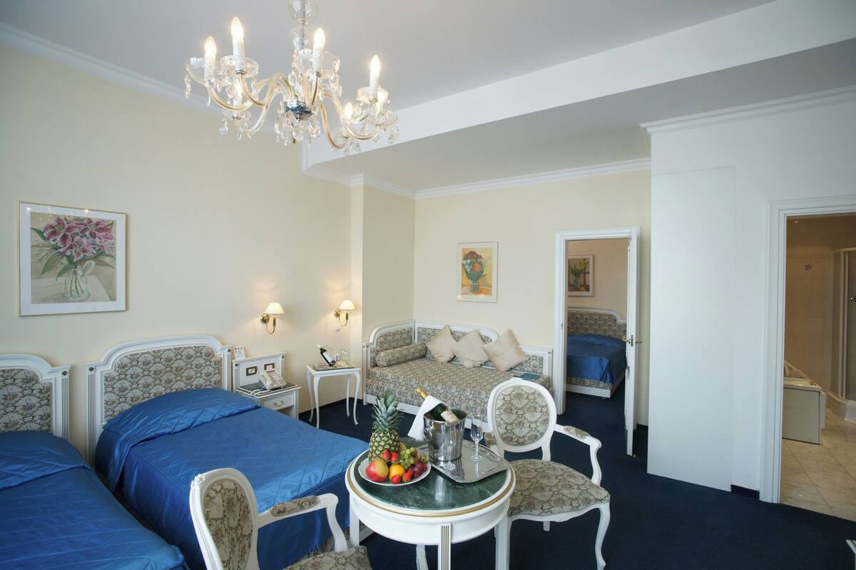 A hotel room with two beds, a dining table with fruit and champagne, a chandelier, and artwork on the walls.