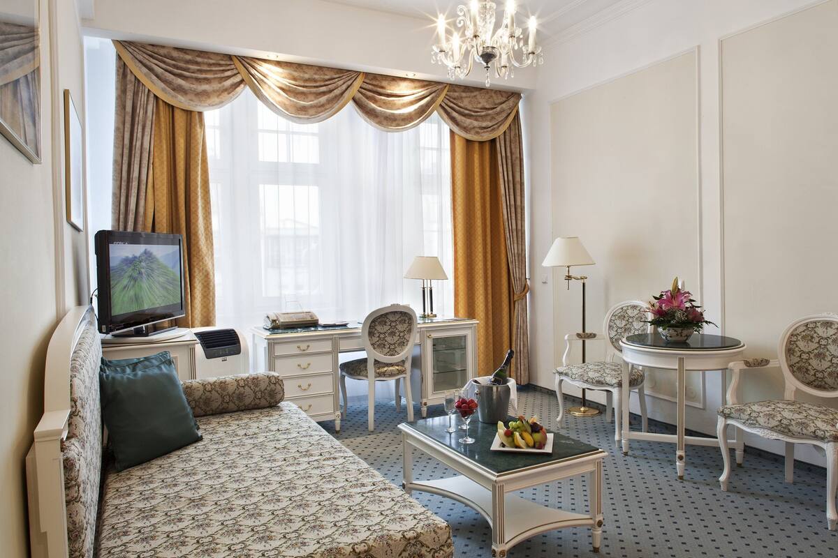 A hotel room with a sofa, a television, a dining table with fruit, two chairs, a lamp, and a desk with a computer.