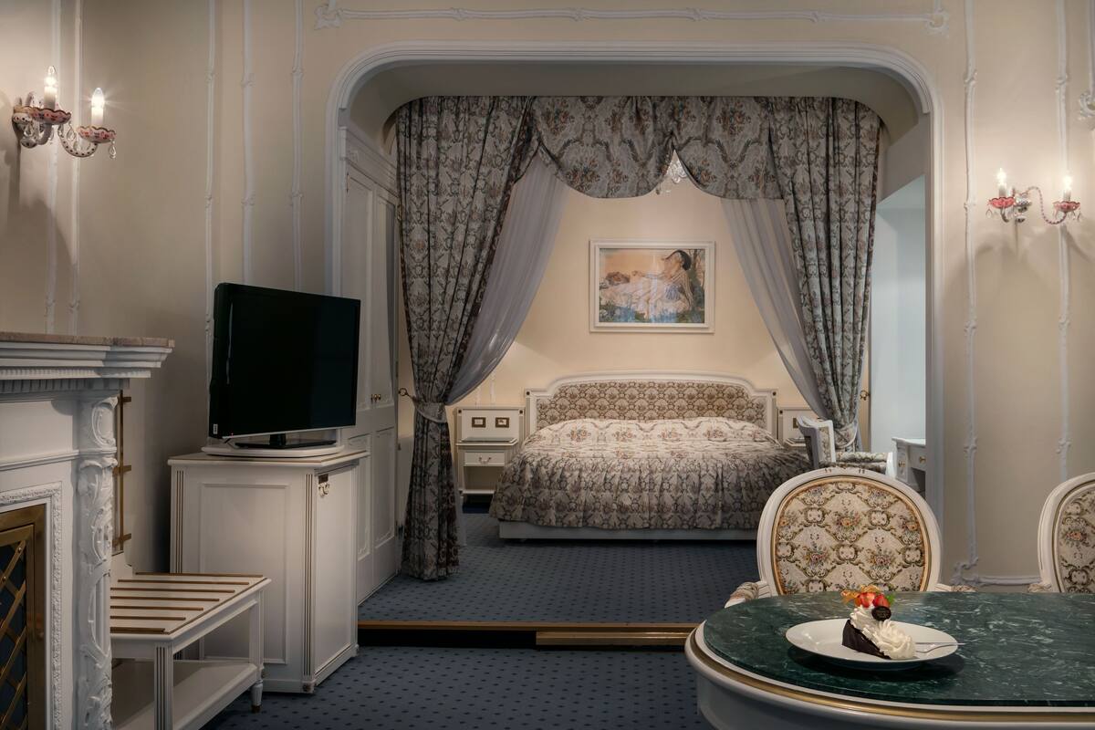 A bedroom with a bed, a television, a dining table with a plate of food, and a painting on the wall.