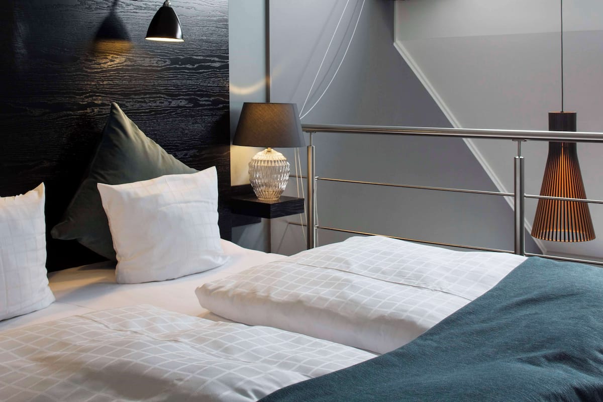 A modern hotel room with a bed, pillows, a nightstand, a lamp, and a view of the outside.