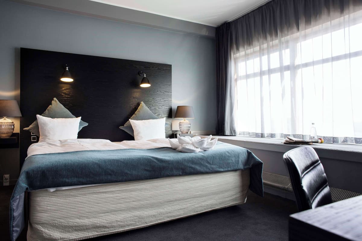 A modern hotel room with a large bed, two bedside lamps, a window with curtains, and a small table with a bottle and glasses.