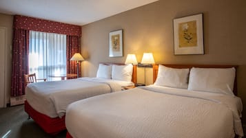 A hotel room with two beds, a chair, a table, and two wall-mounted lamps.