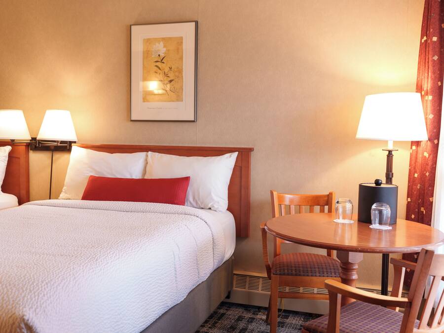 Une chambre d’hôtel avec un lit, deux lampes de chevet, un tableau encadré au mur, une table à manger avec des chaises et une fenêtre avec des rideaux.