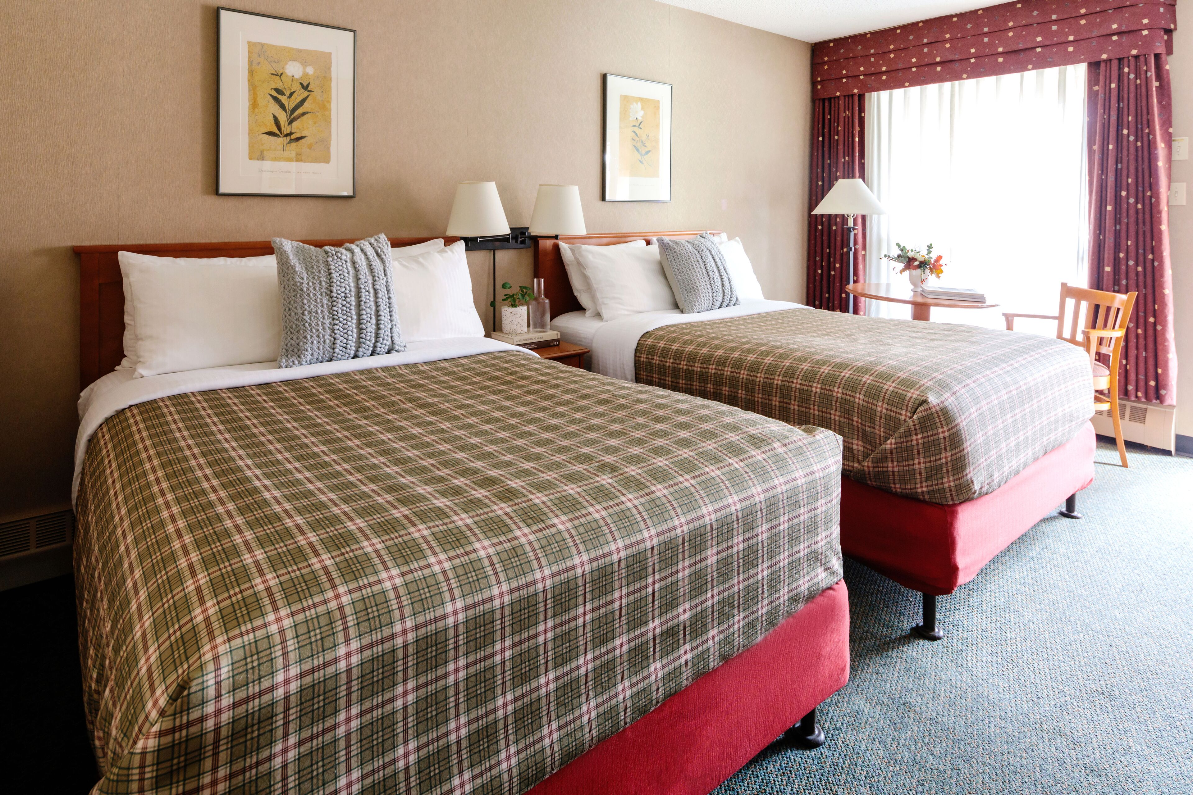 Une chambre d’hôtel avec deux lits, chacun recouvert d’une couverture à carreaux, doté d’une tête de lit en bois et agrémenté d’une table de chevet avec une lampe. On y trouve également une petite table avec un vase de fleurs et une chaise.