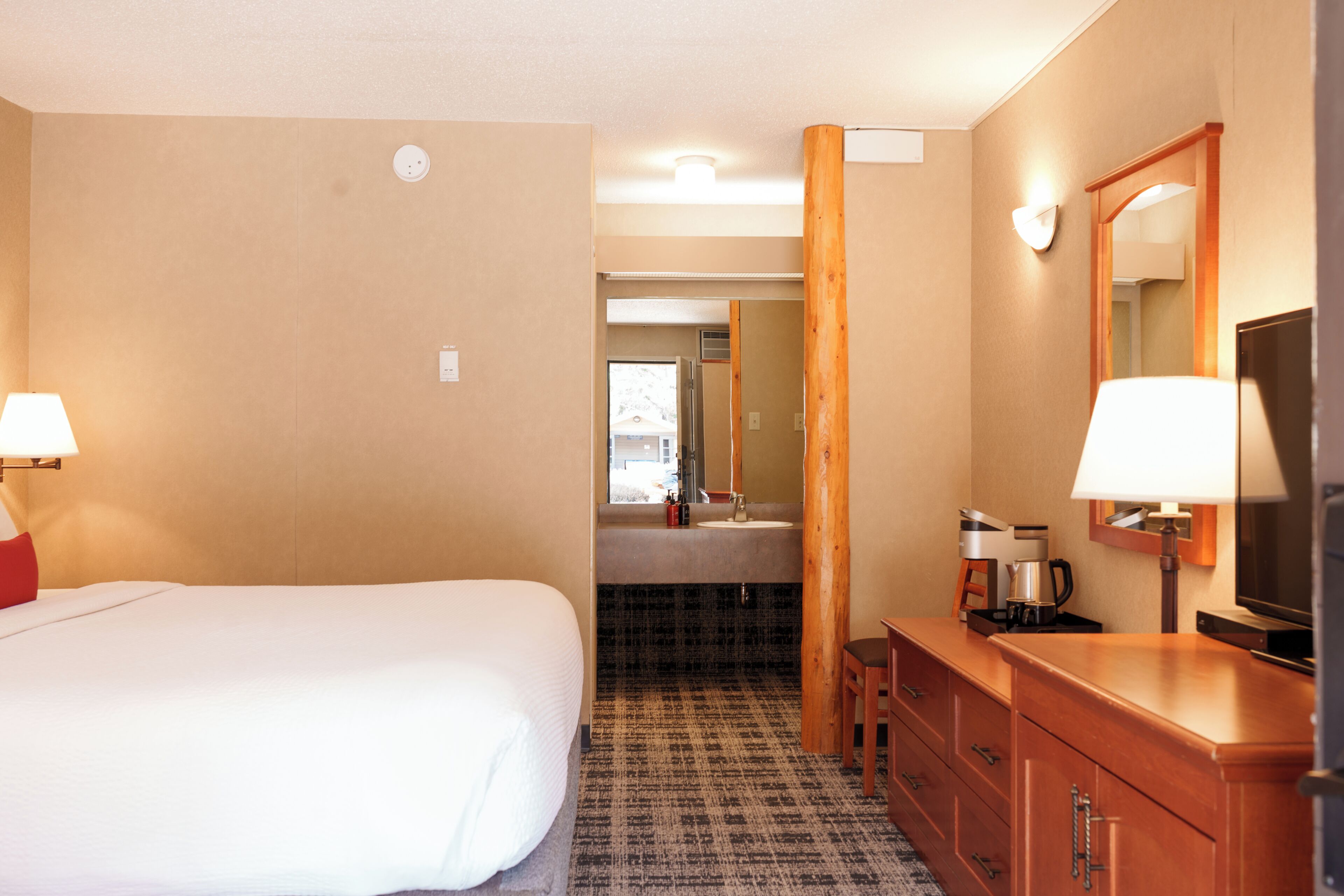 A hotel room with a bed, a desk, a mirror, a coffee maker, and a bathroom visible through an open door.