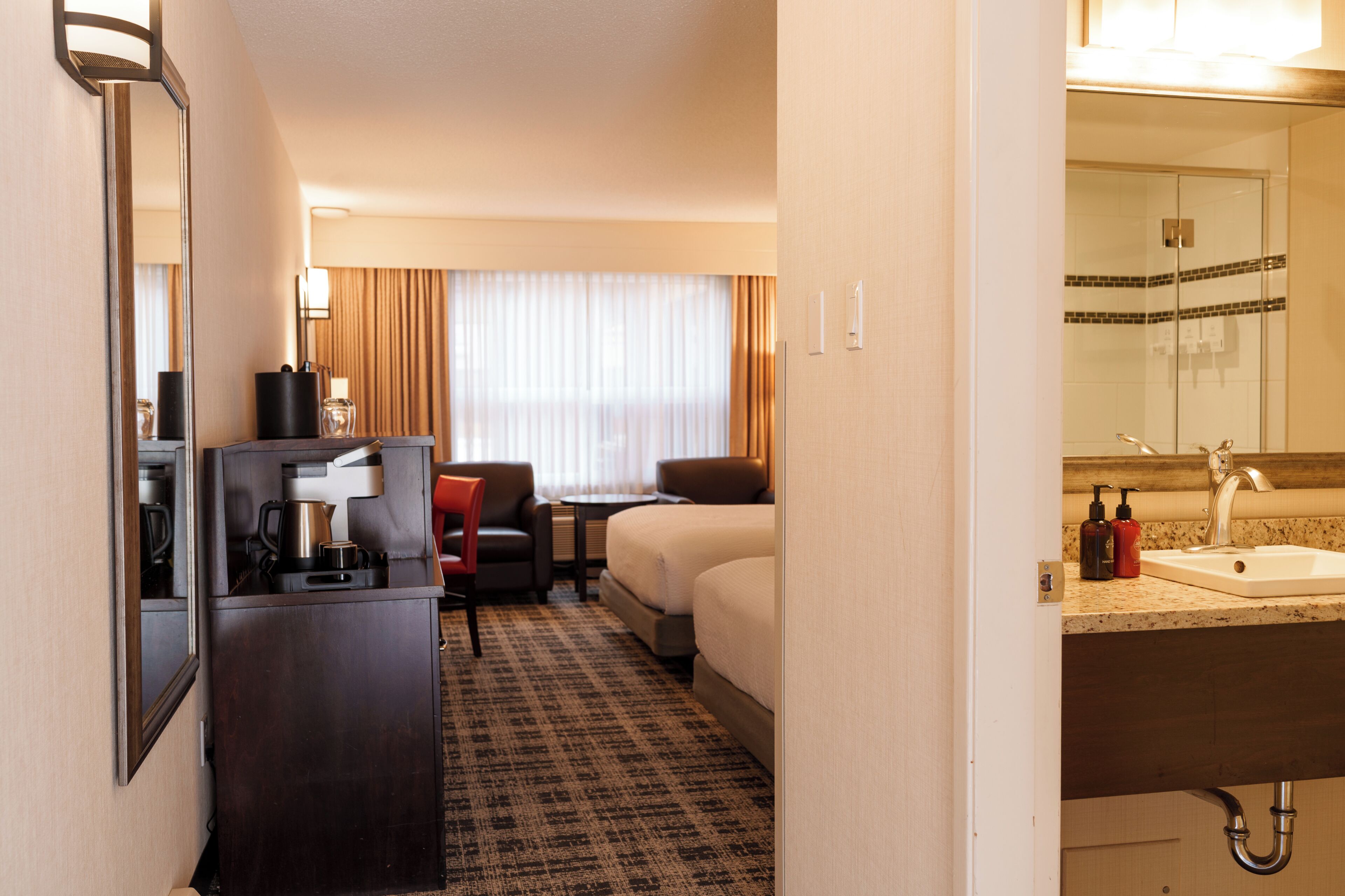 A hotel room with a bed, a desk with a chair, a small table, a mirror, a sink, and a shower area.