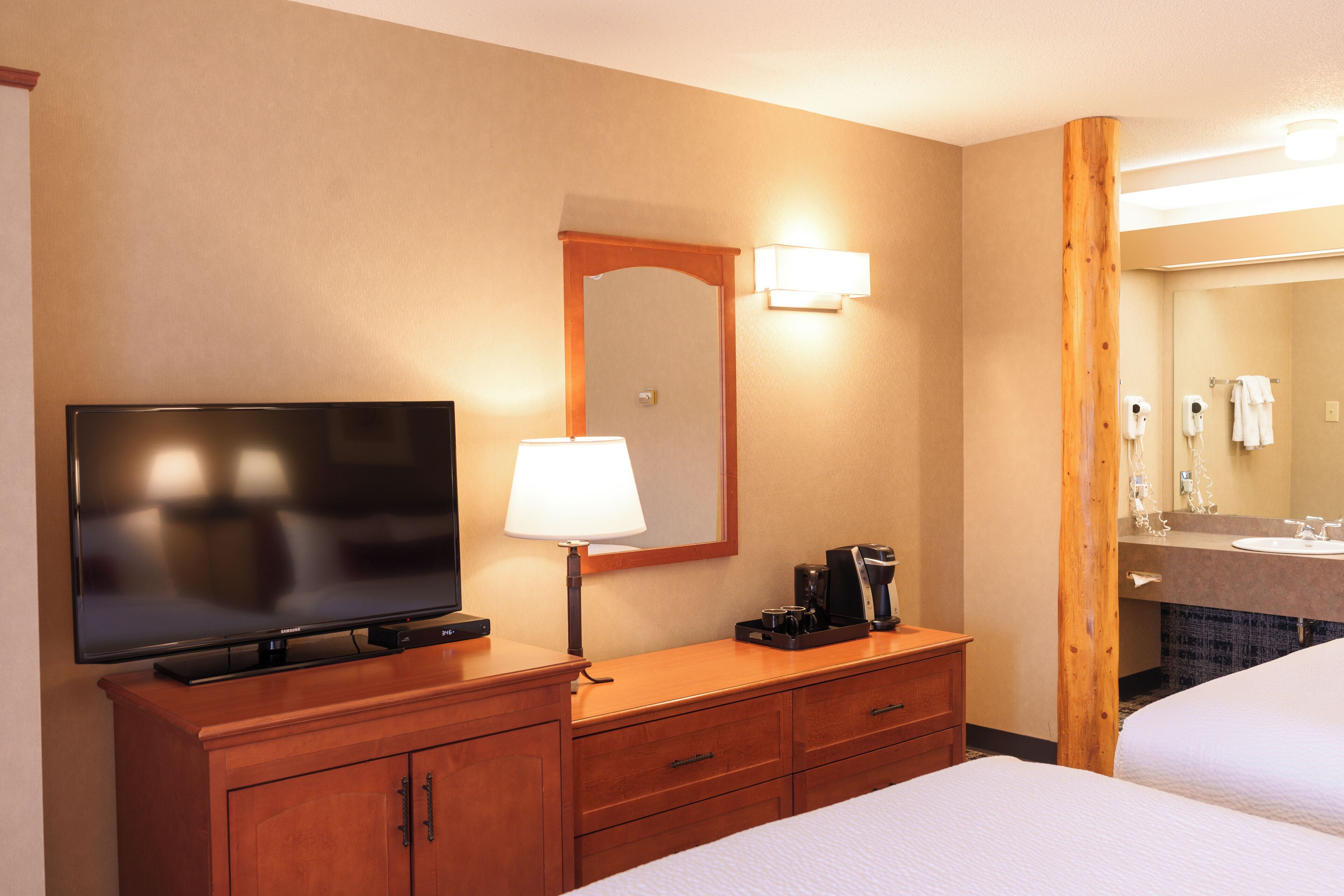 A hotel room with a flat-screen TV, a lamp, a coffee maker, a mirror, and a bed.