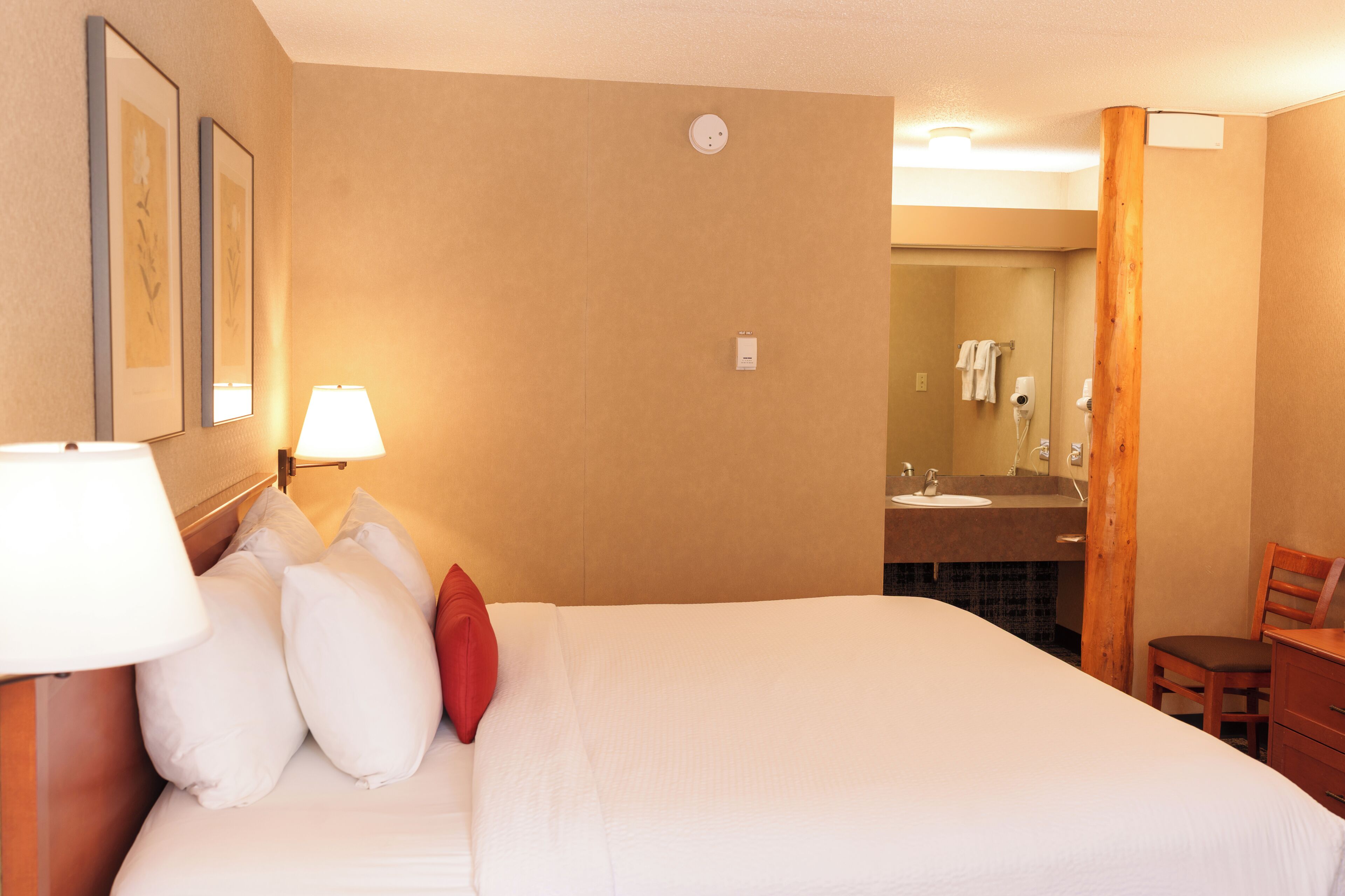 A hotel room with a bed, a desk, a chair, a lamp, a mirror, and a bathroom visible through an open door.