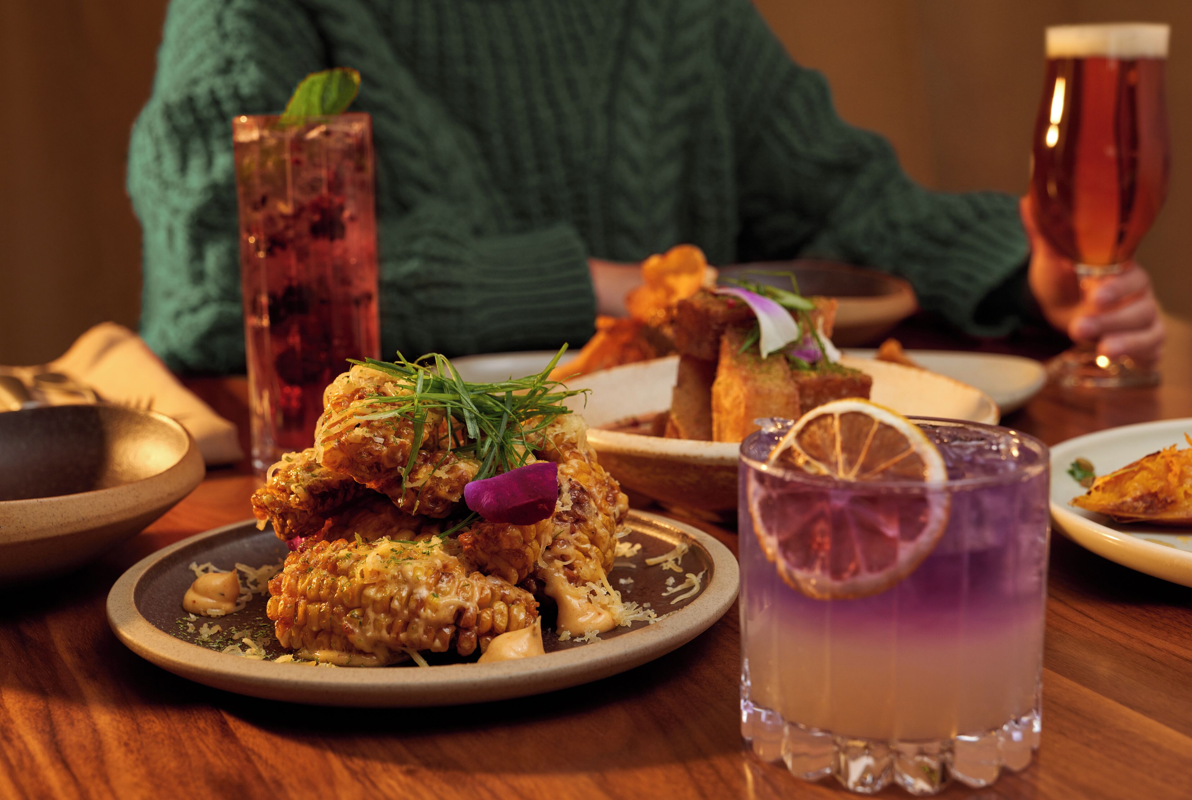 A dining table with various dishes, a tall glass of a purple drink, and a person in a green sweater.