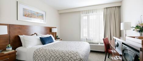 A hotel room with a large bed, a desk with a computer, a chair, a window with curtains, and a framed picture on the wall.