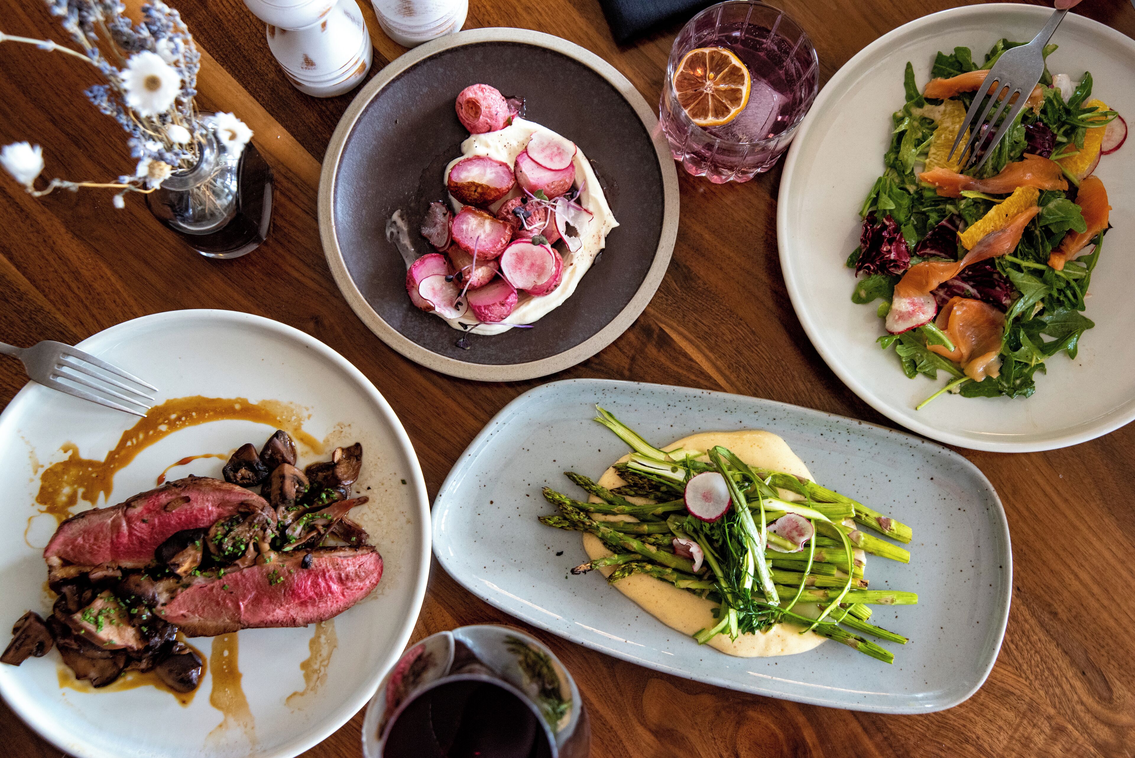 Variedad de platos sobre una mesa de madera, incluyendo un plato con rábanos en rodajas, un plato con espárragos y un plato con carne y champiñones.