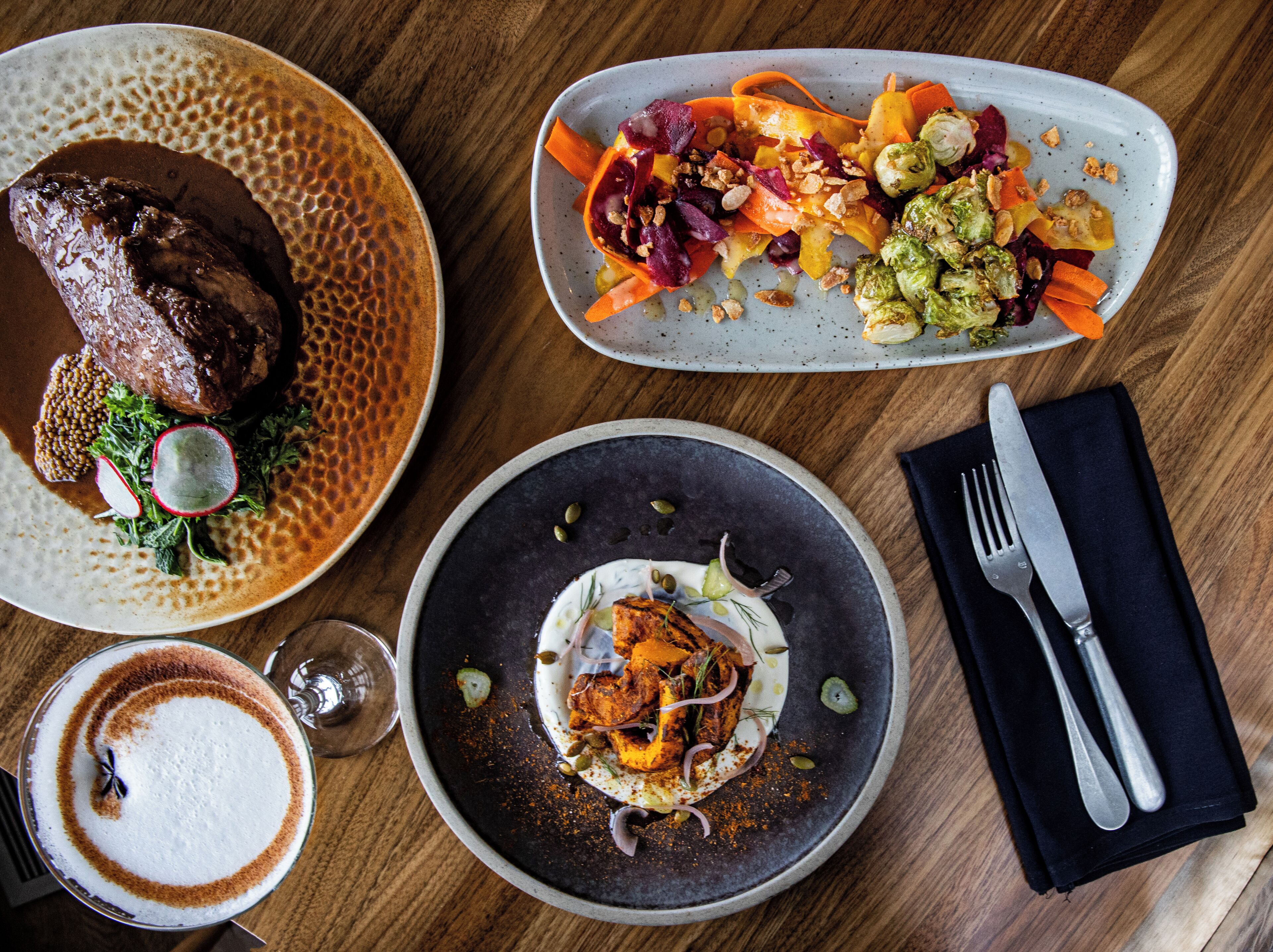 A dining table with three plates of food, a glass of coffee, and cutlery.