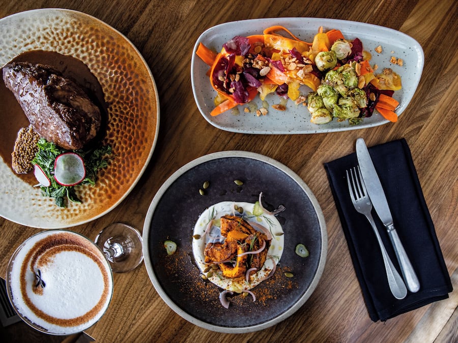 A dining table with three plates of food, a glass of coffee, and cutlery.
