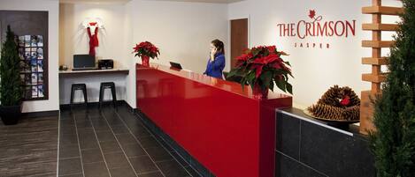 Zona de recepción con mostrador rojo, una recepcionista y dos plantas en macetas con flores rojas.