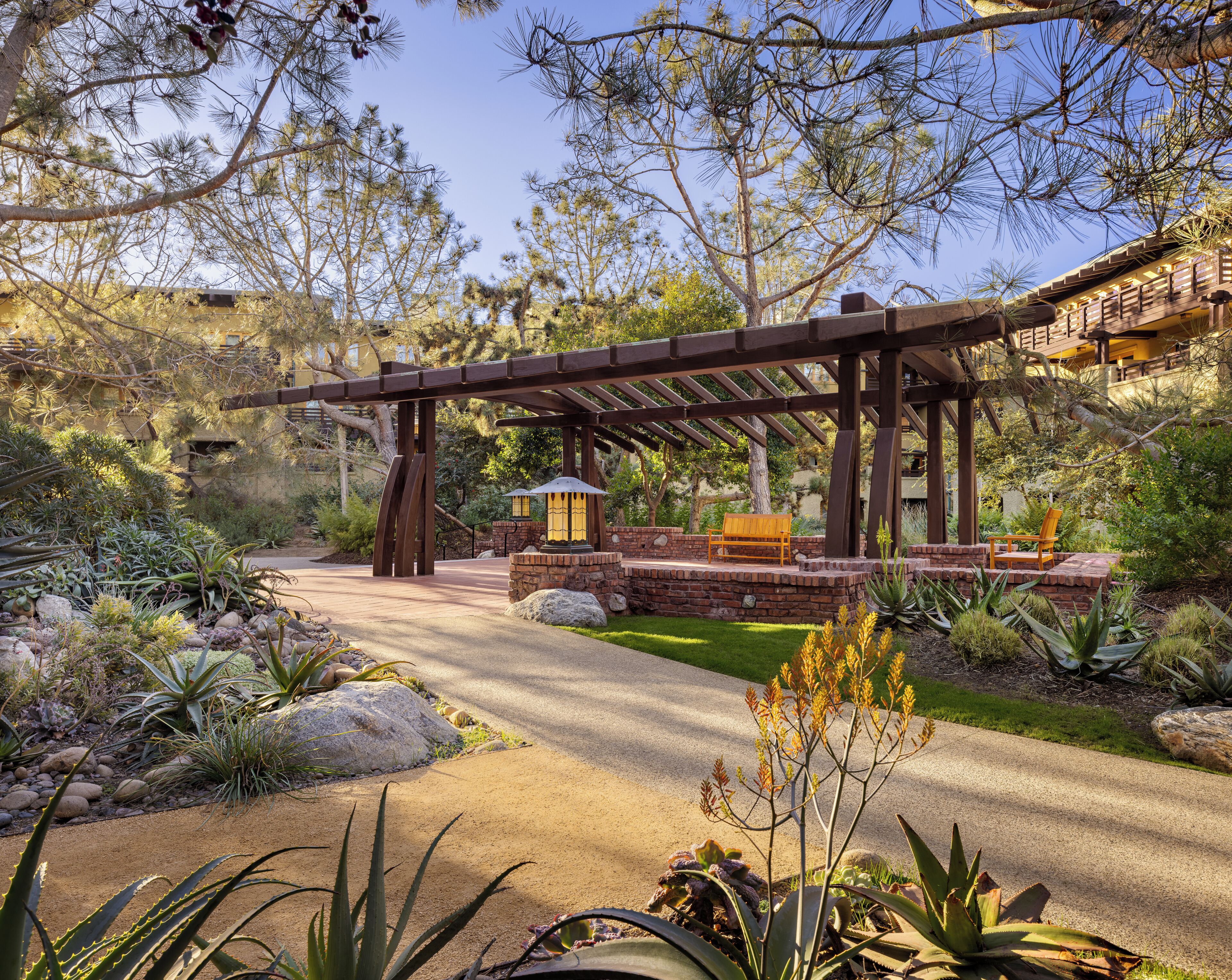 A covered outdoor area with a wooden pergola, a bench, and a pathway leading to a building.