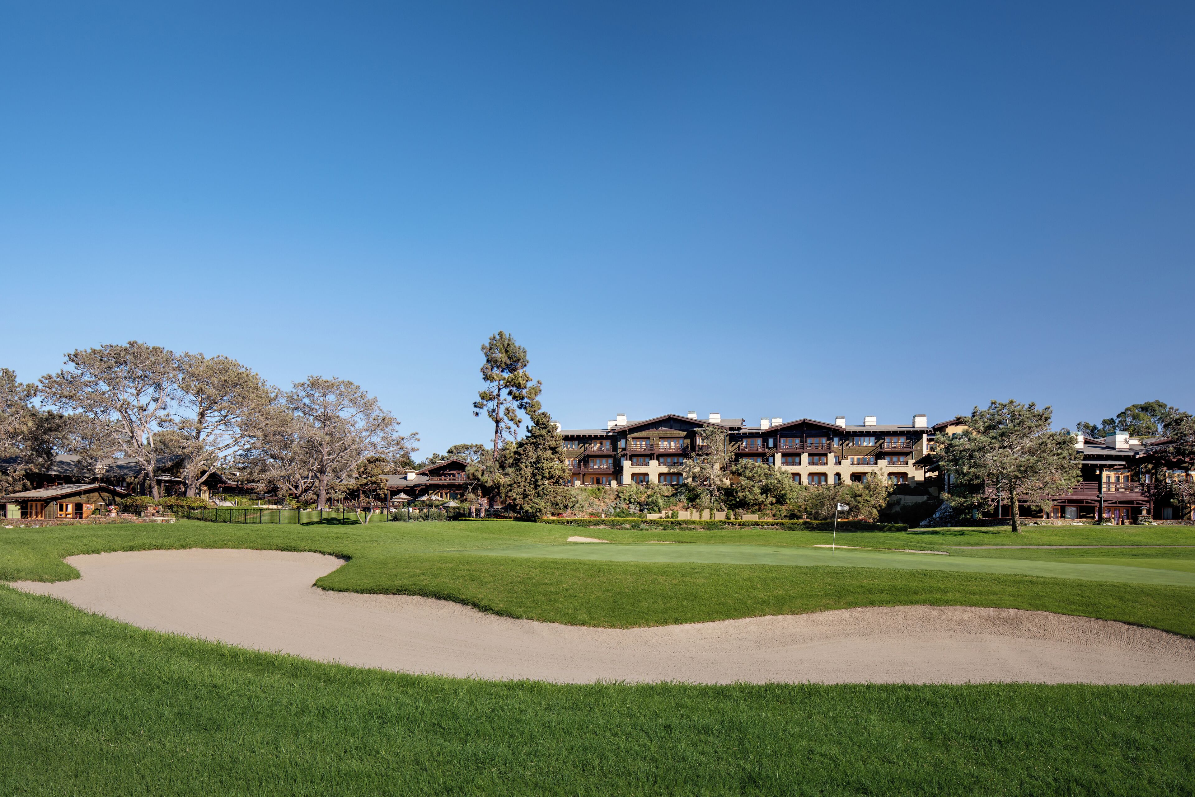 Un campo de golf con un búnker de arena, césped bien cuidado y un fondo de edificios altos bajo un cielo azul despejado.