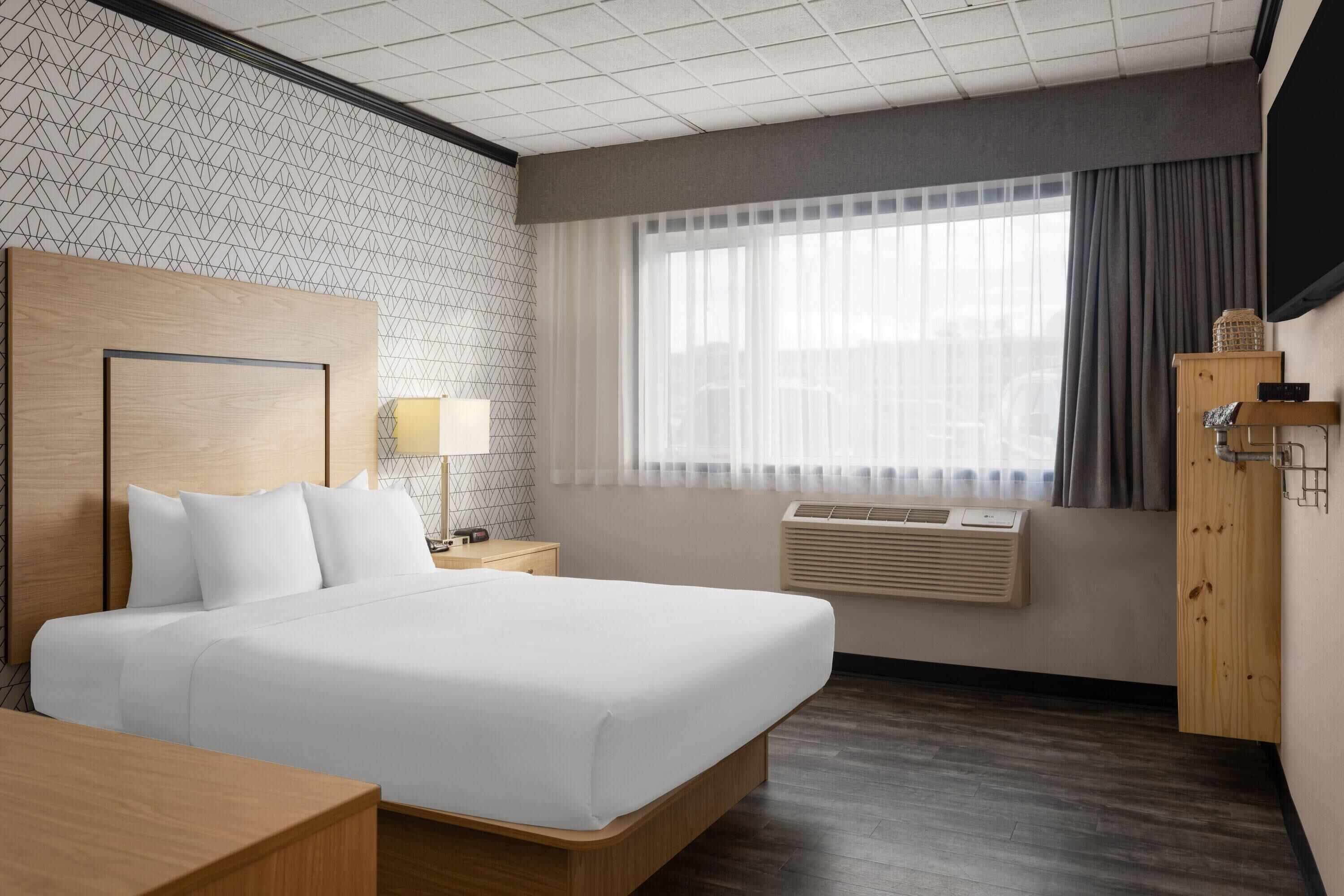 A hotel room with a large bed, a wooden headboard, a bedside table with a lamp, a wardrobe, and a window with curtains.