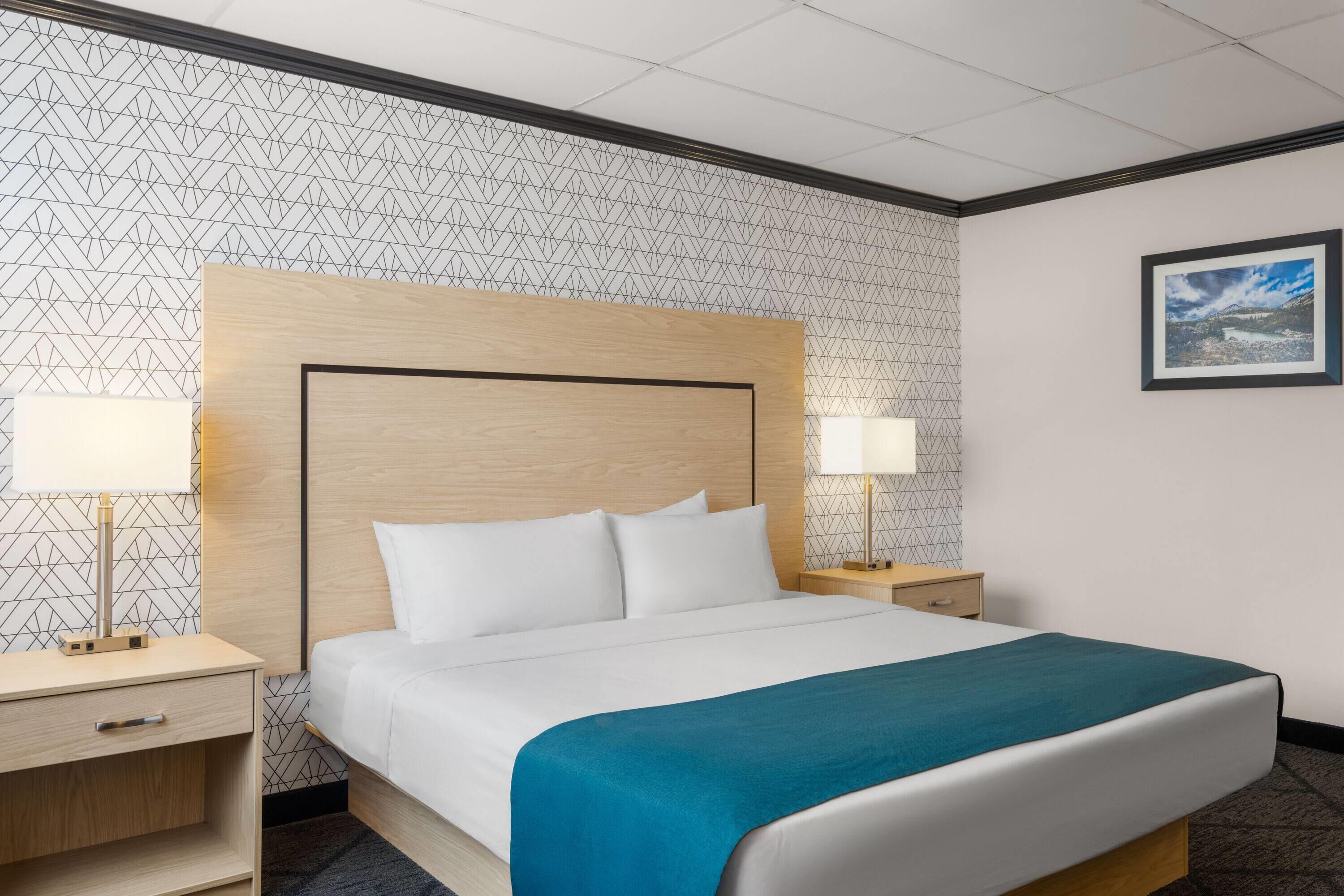 Habitación de hotel con una cama grande, dos mesitas de noche con lámparas, un cabecero de madera y un papel tapiz estampado.