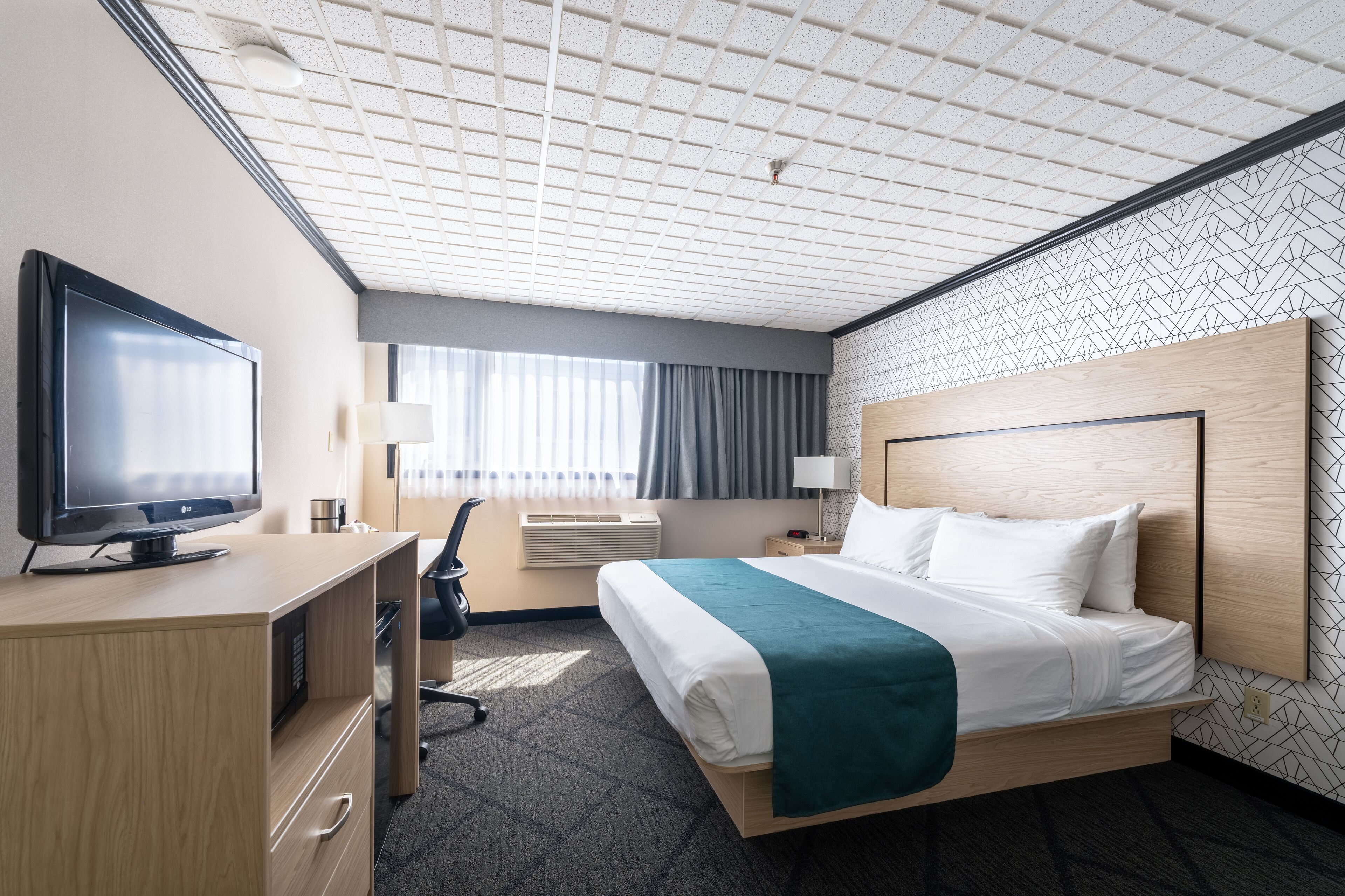 A hotel room with a large bed, a flat-screen TV, a desk with a computer, and a window with curtains.