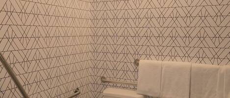 Un baño con pared de diseño geométrico, inodoro blanco, barra de apoyo, toallero y cubo de basura.