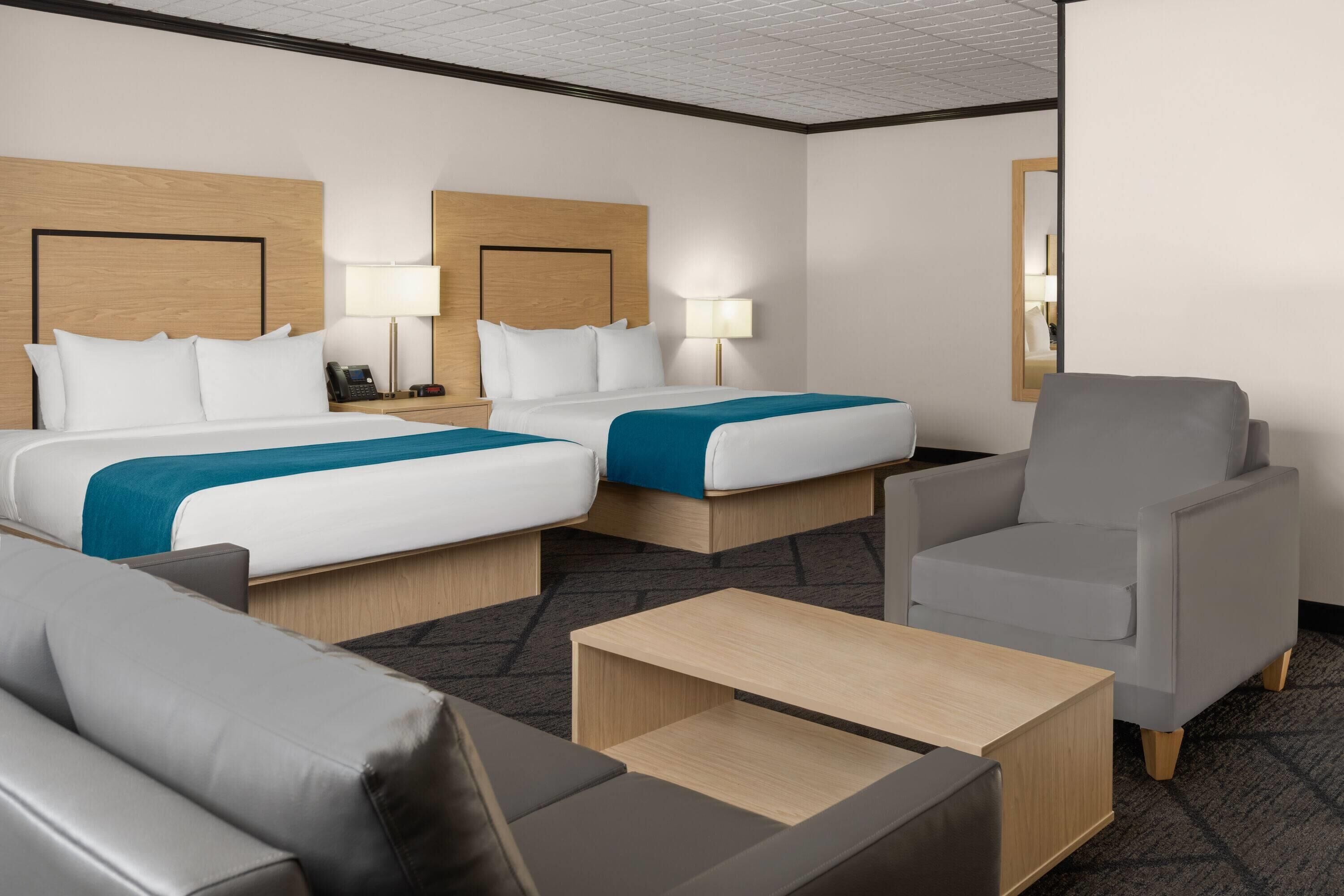 Une chambre d’hôtel avec deux lits, un fauteuil gris, une table basse en bois et une table de chevet avec une lampe.
