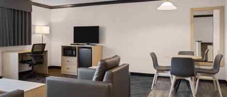 Ein Hotelzimmer mit Flachbildfernseher, Schreibtisch mit Lampe, Sofa, Esstisch mit Stühlen und einem großen Spiegel.