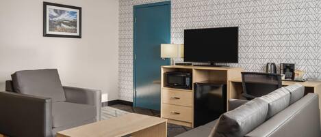 Une chambre d’hôtel moderne équipée d’un canapé, d’un fauteuil, d’une télévision, d’un four à micro-ondes et d’un bureau avec une lampe.