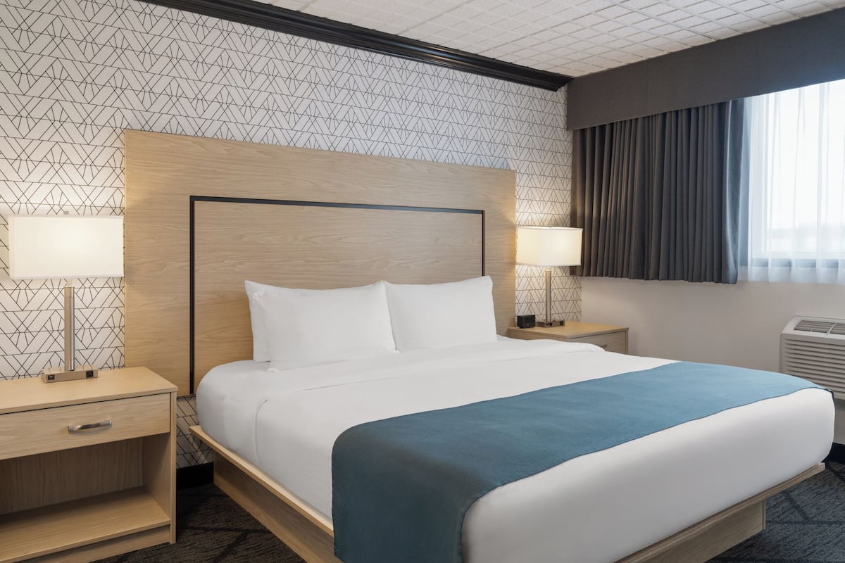 A hotel room with a large bed, two bedside tables with lamps, a headboard, and a window with curtains.