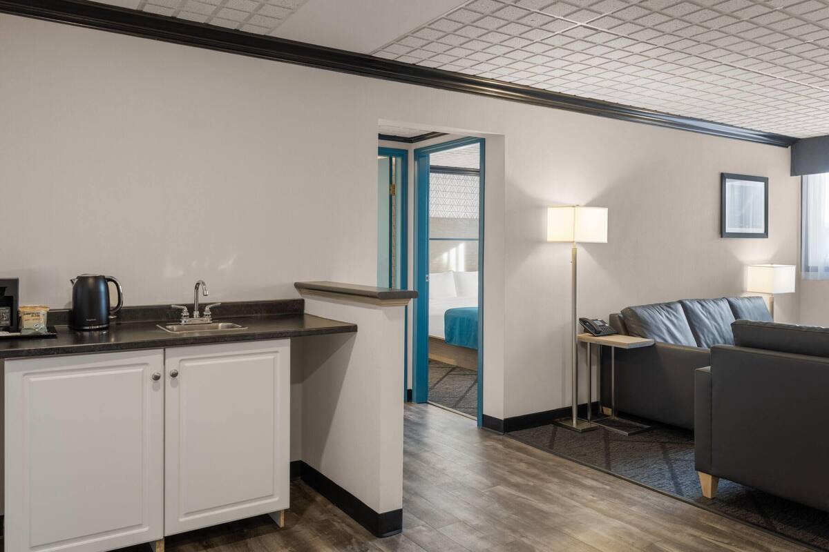A compact hotel room with a kitchenette, a sofa, a bedside table, and a lamp.