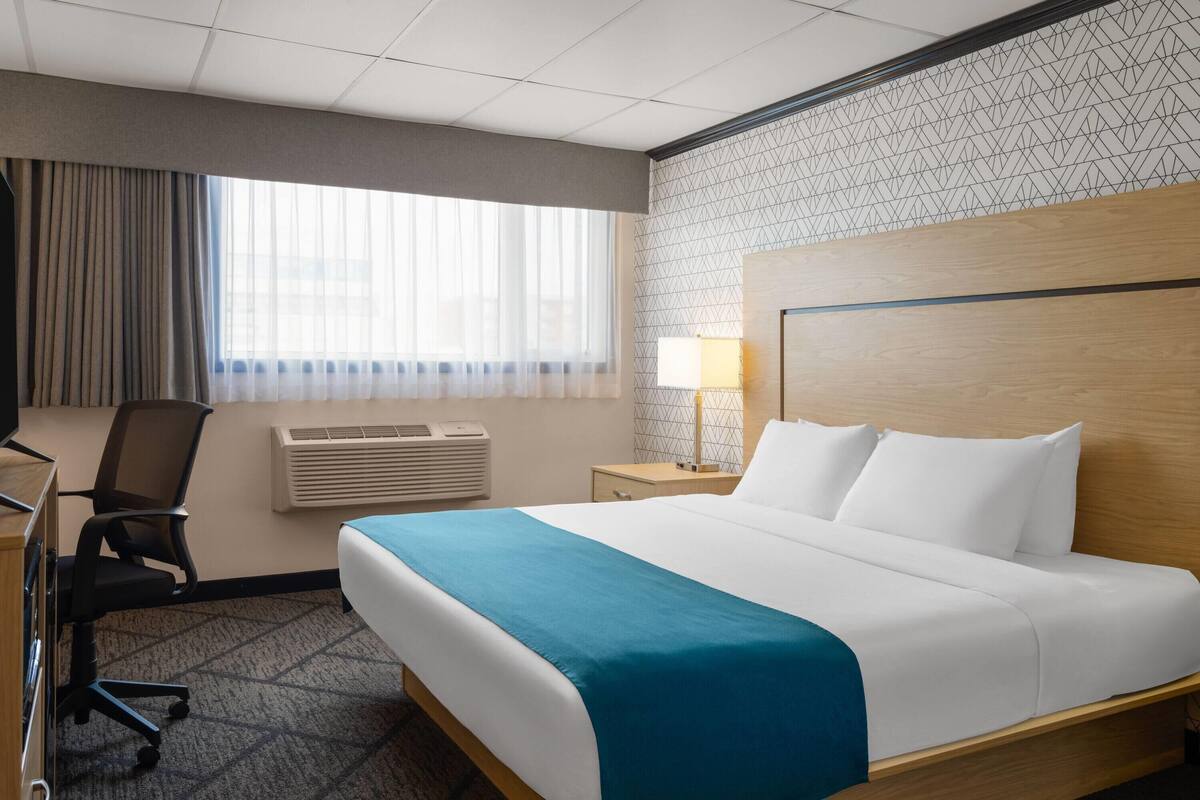 A hotel room with a large bed, a desk with a chair, a lamp, and a window with curtains.
